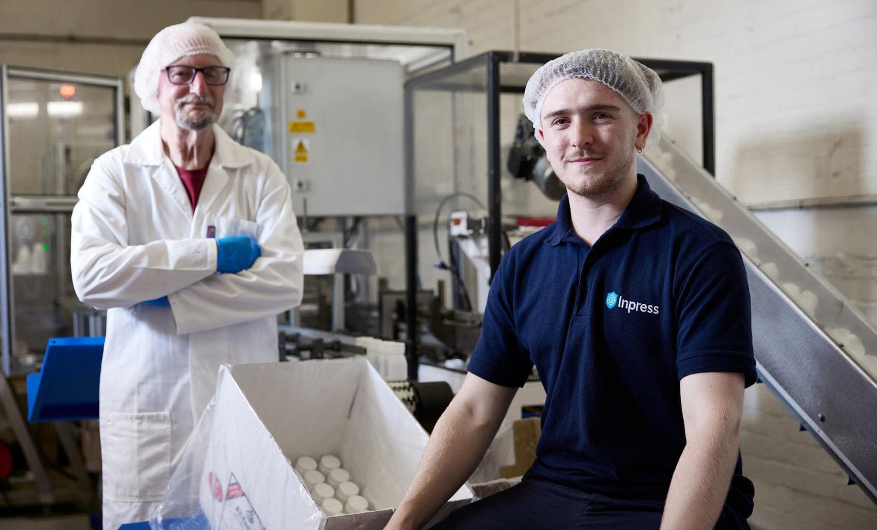 Two Inpress team members in hairnets at a packing line: one in a white lab coat with arms folded, one in a navy Inpress polo shirt seated beside a box of white caps