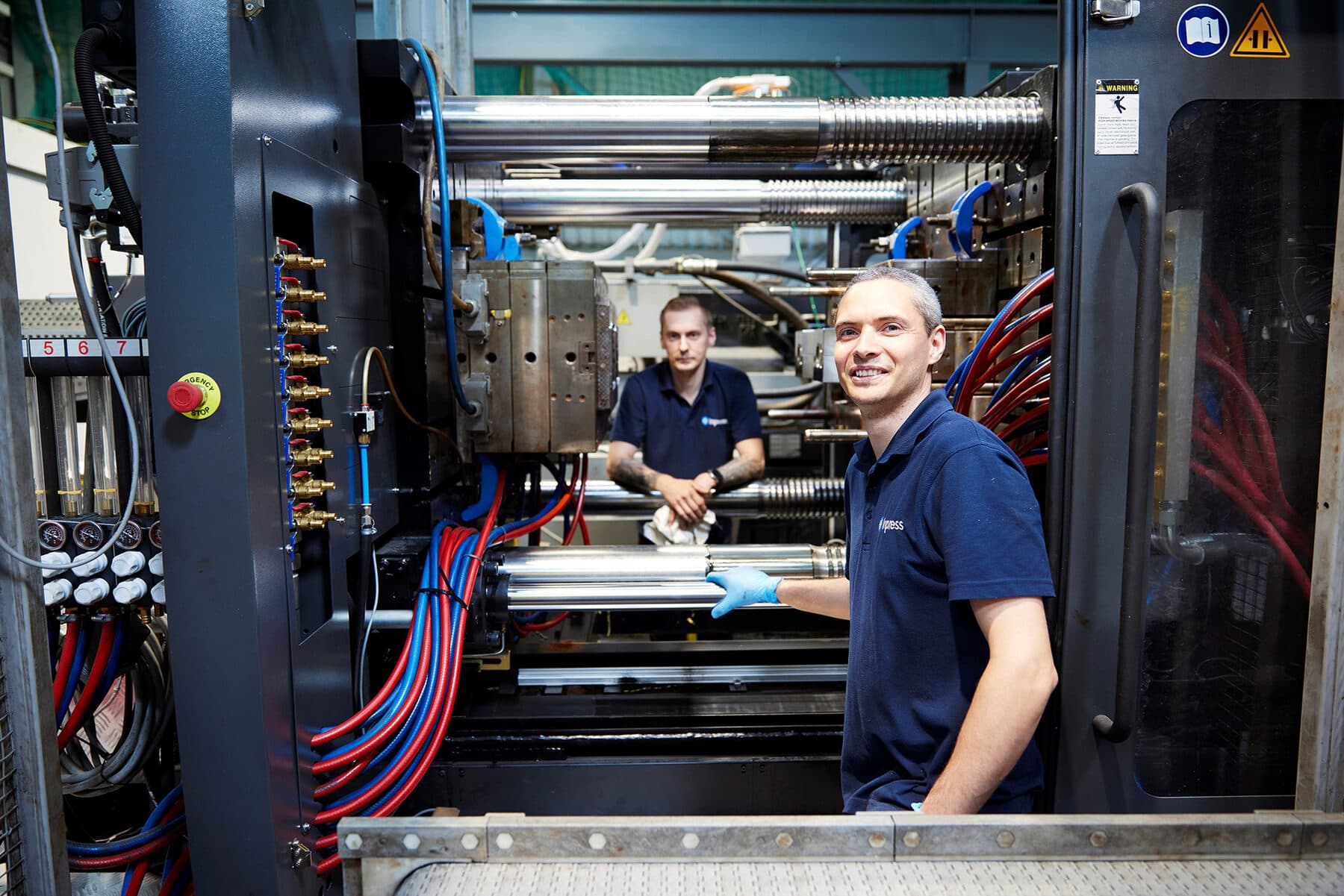 Two Inpress setters in navy polo shirts at an open injection moulding machine, one smiling at the camera