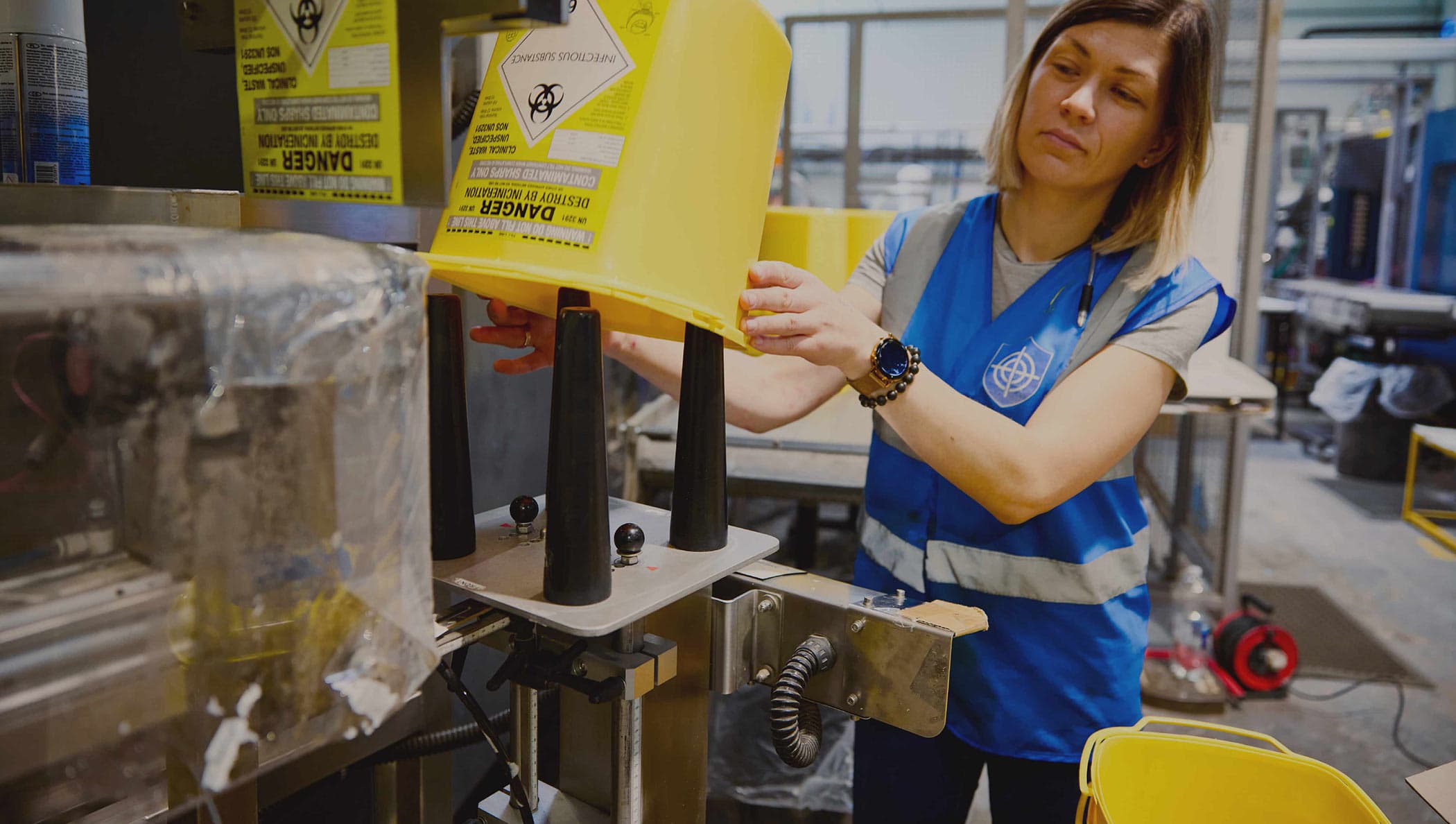 Inpress operator in a blue hi-vis vest placing a yellow sharps container on an assembly fixture