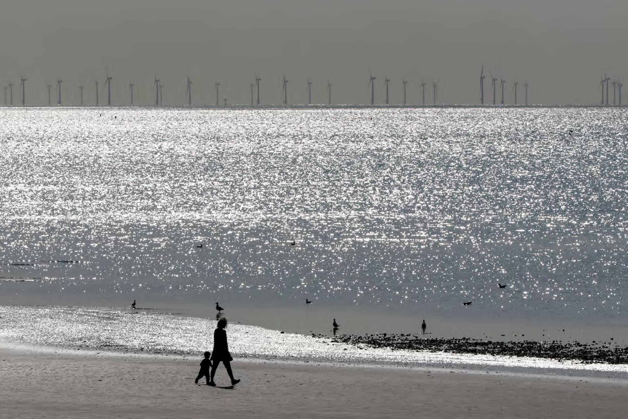 People walking on a beach with offshore wind turbines on the horizon